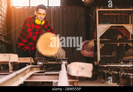 Professional carpenter master in protective glasses and headphones working with cut tree logs to sawmill warehouse. Skilled workman moving wood trunk Stock Photo