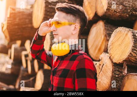 Confident sawmill workman in protective glasses and headphones at wood production warehouse. Young woodworking business owner wearing protective workw Stock Photo