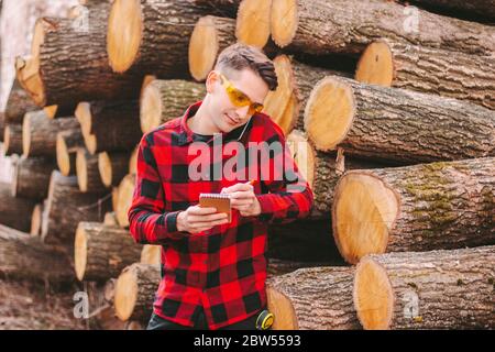 Young workman in protective glasses talking mobile phone and writing to notebook at sawmill factory. Happy wood production mill workman inspect cut tr Stock Photo