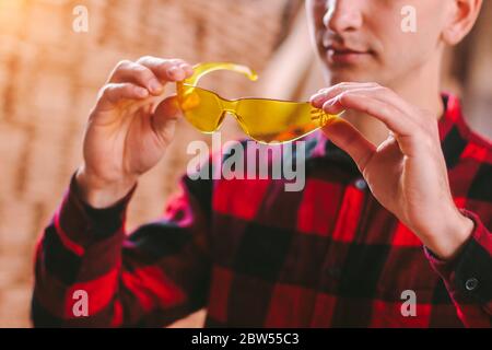 Closeup of man professional carpenter holding protective glasses in hands while working at woodworking workshop. Male cabinet maker wearing eye protec Stock Photo