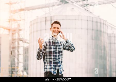 Portrait of young happy business man farmer holding mobile phone in hand and smiling while standing against grain silos warehouse building. Confident Stock Photo