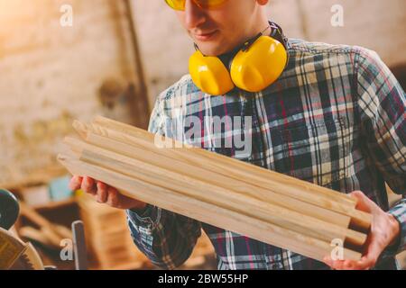 Closeup of young male carpenter with protective headphones on neck holding stack of wooden planks in hands while working at sawmill. Professional arti Stock Photo