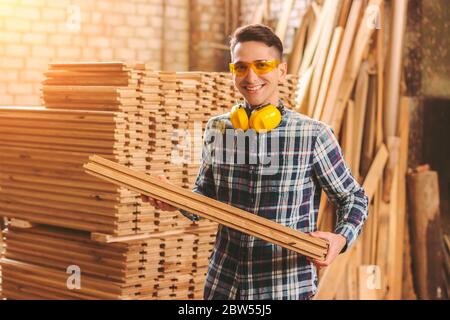 Happy man cabinet maker in protective glasses and headphones holds pile of wooden material in hands and smiling at sawmill factory. Male carpenter woo Stock Photo