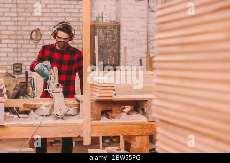 Skilled cabinet maker in protective headphones, glasses and mask using circular saw machine for cutting wooden boards at workshop. Professional carpen Stock Photo