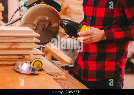 Closeup of professional cabinet maker holding yellow protective headphones in hands while working at sawmill. Skilled carpenter workplace for woodwork Stock Photo