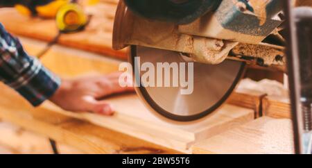 Closeup banner of skilled caprenter using electric circular saw for cutting wood board at sawmill. Professional joiner cutting wooden plank with circu Stock Photo