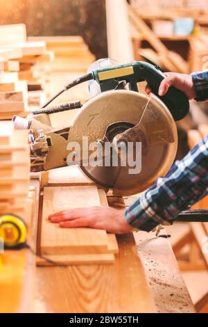 Closeup skilled man hands holding electric circular saw for cutting wood board at woodworking workshop. Professional carpenter using circular saw at s Stock Photo