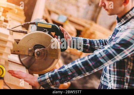 Closeup professional carpenter hold electric circular saw at sawmill. Skilled joiner using circular saw for cutting wood board at woodworking workshop Stock Photo