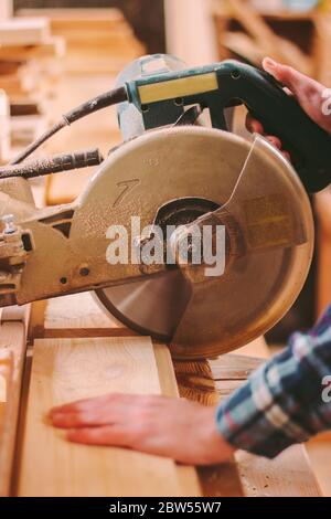 Closeup of skilled cabinet maker using electrical circular saw at woodworking workshop. Professional carpenter cutting wood board with circular saw at Stock Photo