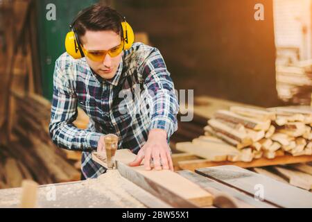 Professional carpenter in protective glasses and headphones using electric circular saw at woodworking workshop. Skilled cabinet maker working with sa Stock Photo