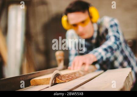 Professional carpenter using sawing machine for cutting wooden board at sawmill. Skilled cabinet maker working with electric circular saw at woodworki Stock Photo