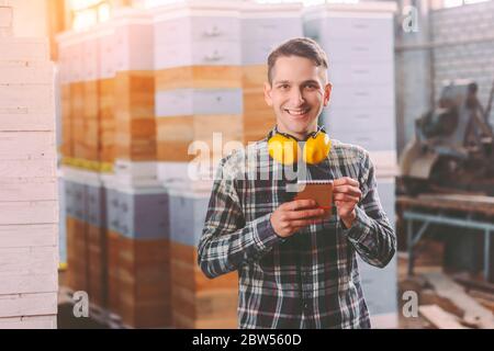 Happy man supervisor with protective headphones on neck writing in notebook while inspecting wooden material at sawmill. Young warehouse worker at lum Stock Photo