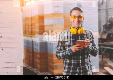Happy sawmill business owner in protective glasses and headphones writing in notebook while supervising storage inventory. Young warehouse worker insp Stock Photo