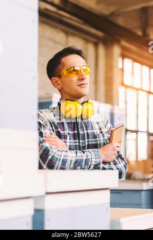 Young man small sawmill factory owner in protective glasses and headphones dreaming with crossed arms about big future for his business and smiling. W Stock Photo
