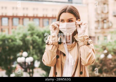 Young happy elegant girl in medical gloves put on protective face mask outdoors. Beautiful young hipster woman in protective gloves wear medical face Stock Photo