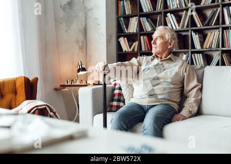 Senior man alone sitting at library holding walking stick looking out thoughtful Stock Photo