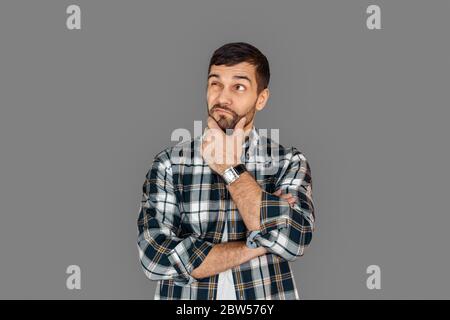 Freestyle. Young man in checkered shirt standing isolated on grey crossed arms touching chin thoughtful Stock Photo