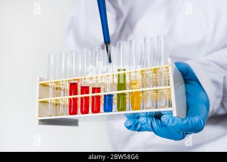 Researcher dripping blue fluid into glass test tubes in plastic rack by pipette. Scientist in protective wear and glove holding in hand lab equipment. Stock Photo