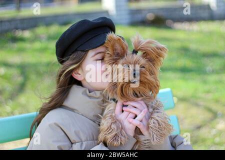 A girl in a black cap and jacket kisses her Yorkshire terrier, which she holds in her arms while walking in the park Stock Photo