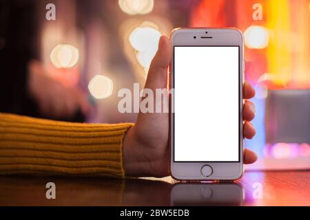 Mockup image of a hand holding a white mobile phone with a blank white screen. blurred background with colored bokeh on a table in a cafe. selective focus Stock Photo