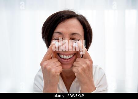 Smiling senior woman applying anti-aging face cream at home. Happy mature woman using day moisturizer Stock Photo