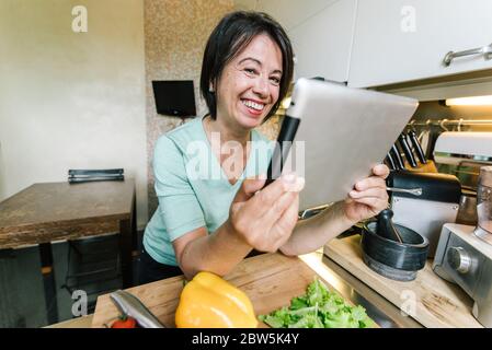 Older senior woman chef cooking in the kitchen at home reading recipe on a tablet device. Stock Photo