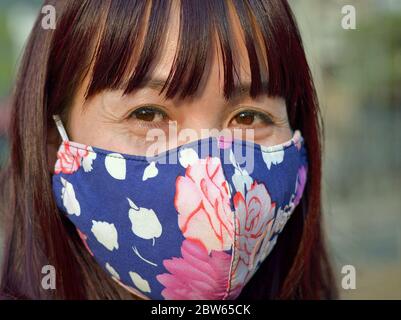 Mature Asian woman with beautiful brown eyes covers her nose and mouth with a washable ear-loop face mask. Stock Photo