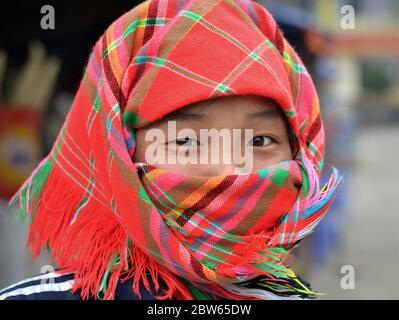 Vietnamese H'mong hill-tribe girl with smiling Asian eyes covers her nose and mouth with a red-checkered headscarf. Stock Photo