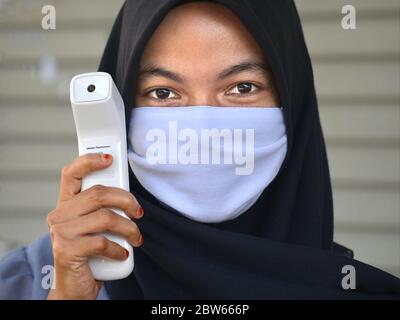 Malay girl wears a black traditional hijab with mouth-covering veil and poses for the camera with a modern infrared thermometer. Stock Photo