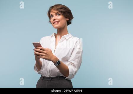 Young beautiful woman with dark short hair in white shirt holding cellphone in hands while dreamily looking aside over blue background Stock Photo