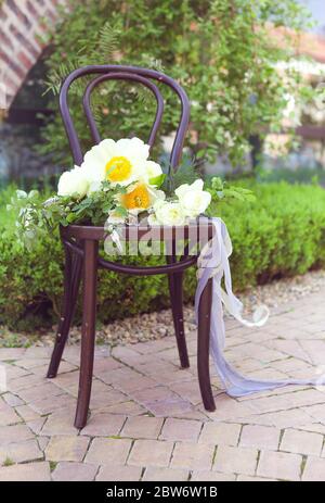 Elegant bouquet of white flowers with decorative ribbons arranged on retro wooden chair placed on paved terrace in summer garden Stock Photo