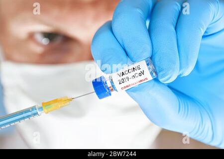 Man hand in blue nitrile glove hold small vial with coronavirus vaccine, to inject it with green orange syringe, blurred face with white virus mask in Stock Photo