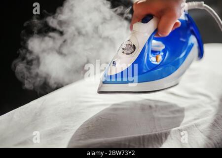 A man holds a new blue modern iron in his hand, from which clouds of steam are coming, and is about to stroke a crumpled cloth in the sunlight. Stock Photo