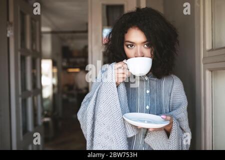Nice African American girl drinking coffee wrapped in plaid. Beautiful lady with dark curly hair standing with cup of coffee in restaurant. Portrait Stock Photo
