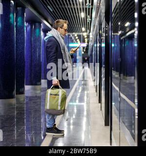 Caucasian young man with backpack using cellphone, waiting for the train at subway station platform early morning, holding a cup of coffee, going to w Stock Photo