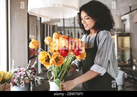 Beautiful florist in apron creating bouquet of colorful tulips. Pretty African American girl with dark curly hair working with flowers Stock Photo