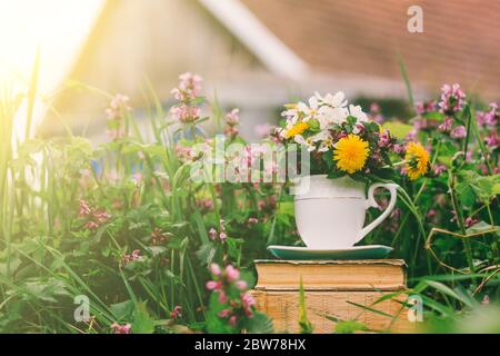 rural landscape a stack of old books with a glass of flowers on a background of flowering grass and a rustic house on a spring or summer sunny morning Stock Photo