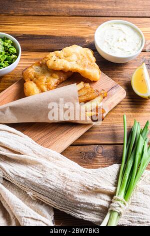 Traditional Fish and chips with mashed peas, tartar sauce inn crumpled paper cone on wood chopping board dip and lemon - fried cod, french fries, lemo Stock Photo