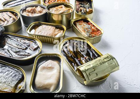 Opened preserve cans with Saury, mackerel, sprats, sardines, pilchard, squid, tuna over white textured stone table side view space for text. Stock Photo