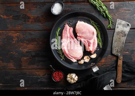 Raw pork meat chopes with herbs in iron skillet with burcher cleaver knife for chopping in butchery on a dark wood surface and ingredients for cooking Stock Photo