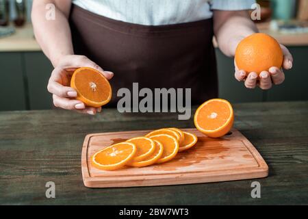 Hands of contemporary housewife in apron standing by kitchen table and holding fresh oranges over wooden board with juicy slices Stock Photo