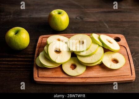Pile of fresh green apples ready to be put on fruits dryer on wooden cutting board and two granny smiths near by on kitchen table Stock Photo