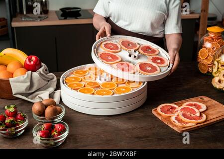 Contemporary housewife standing by kitchen table and putting fruit dryer tray with sliced grapefruit on that with orange slices Stock Photo