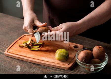 Hands of housewife peeling fresh kiwi over chopping board by kitchen table while preparing them for slicing and drying for winter Stock Photo