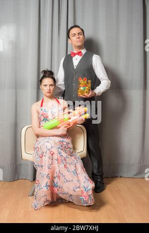 Eccentric marriage portrait with solemn attitude. The woman, seated in an elegant flowered dress, carrying a toy weapon. The man, standing, wearing an Stock Photo