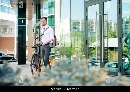 Contemporary young office manager in formalwear standing by door of business center while going to go back home by bicycle Stock Photo