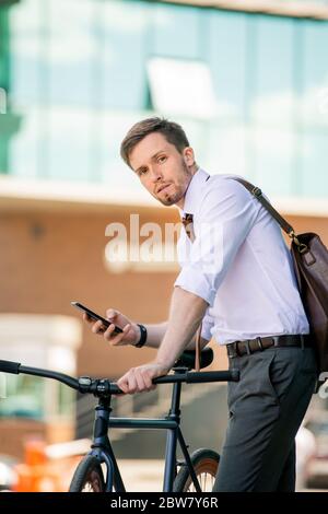 Young man with bicycle and smartphone looking at you while standing in front of camera on background of modern business center Stock Photo