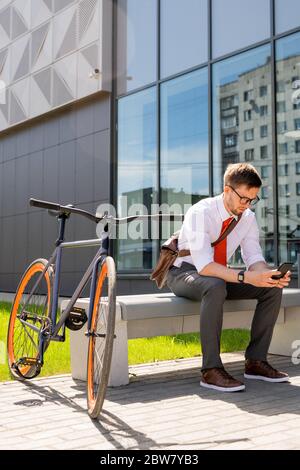 Young businessman sitting on bench on sunny summer day and scrolling in smartphone while having break outdoors by business center Stock Photo