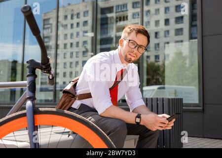 Young businessman looking at you while sitting on bench and using smartphone in urban environment after working day Stock Photo