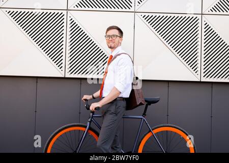 Happy young elegant man standing by wall of modern architecture while going to use bicycle to go home or to cafe after working day Stock Photo